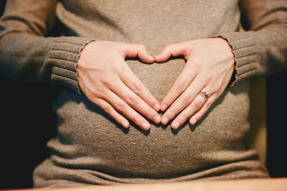 Close-up of a pregnant person wearing a soft brown sweater, gently forming a heart shape with their hands over their belly, symbolizing love, connection, and care during pregnancy.