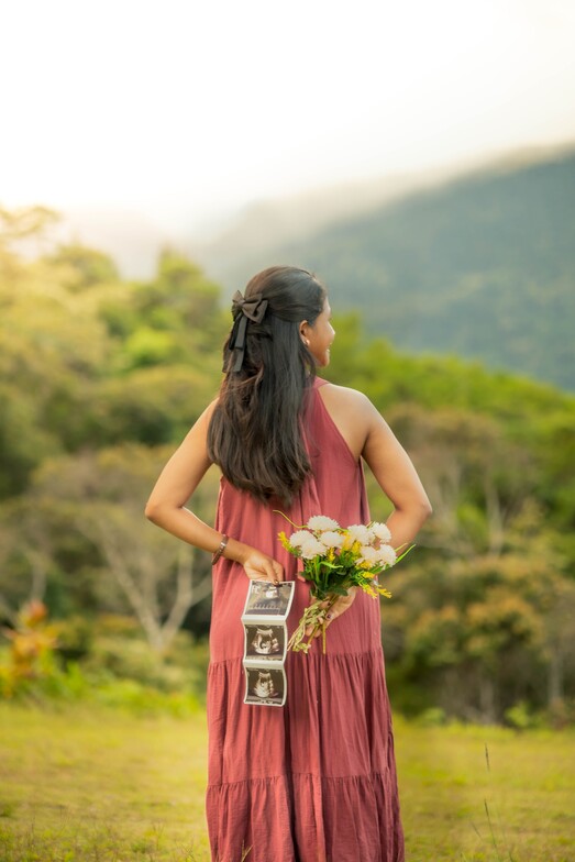 Woman in a rose-colored dress holding ultrasound photos and white flowers outdoors.